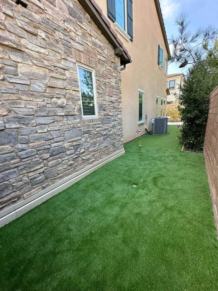 Side yard with artificial green grass, reminiscent of a putting green installation, bordered by a stone wall on one side and a beige stucco wall with windows on the other. An HVAC unit is situated near the house's back wall, and a small American flag decoration is planted in the lawn.