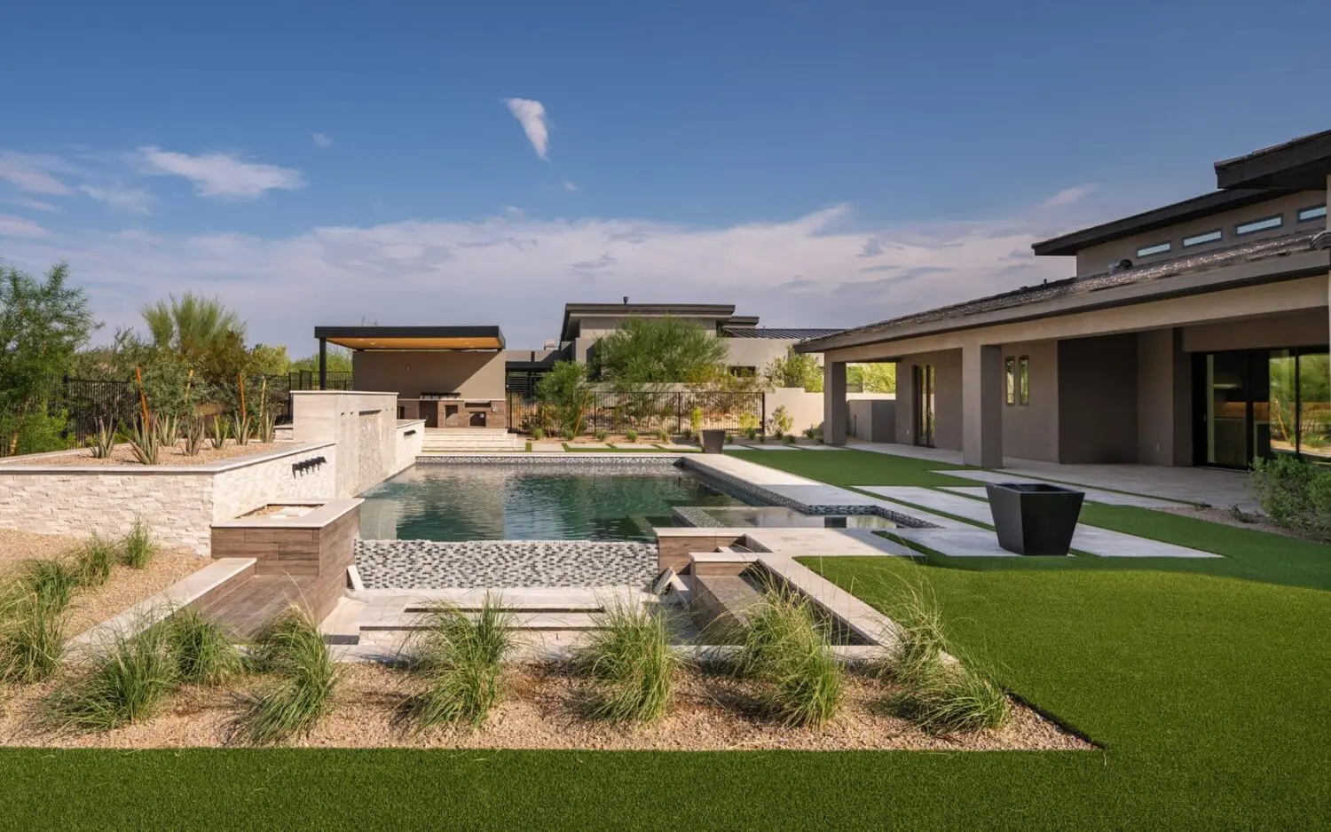 Modern outdoor area with sleek, geometric pool and spa, surrounded by well-manicured lawns and landscaping. This stunning outdoor space features a pergola, contemporary patio furniture, and a large grassy area with pathways. The surroundings are accented by desert plants and clear skies in Surprise AZ.