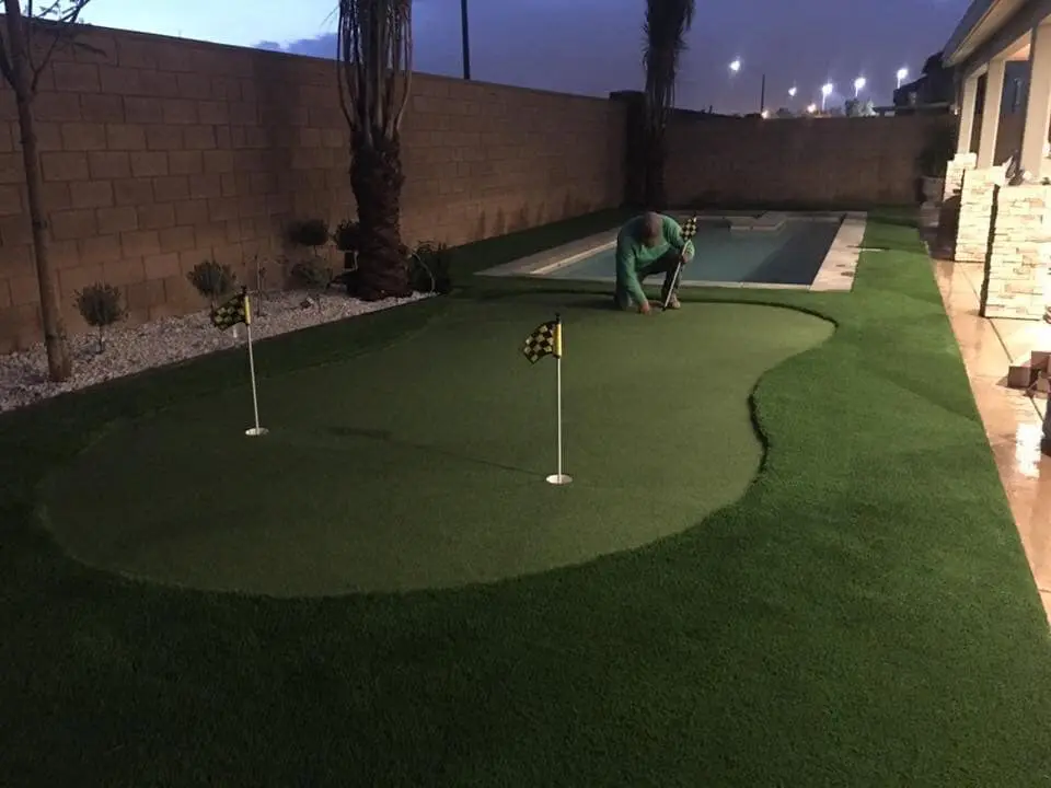 A person is setting up a backyard golf green with expert installation next to a rectangular swimming pool. The green, covered in artificial grass, has two small flags with checkered black and yellow patterns. The scene is lit by outdoor lights in the evening, with a brick wall and plants bordering the area.