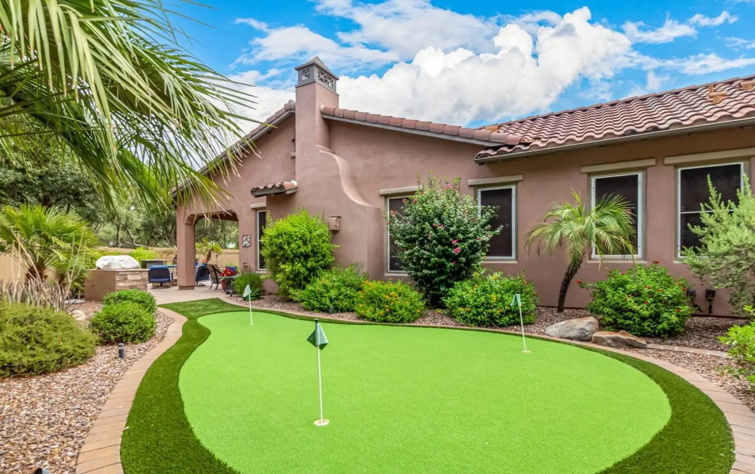 A small backyard putting green with precisely trimmed artificial grass, expertly installed by contractors in Peoria, Arizona, surrounded by lush green shrubs and plants. There's a ceramic pot with pebbles in the forefront and various other plants adding to the cozy garden ambiance.