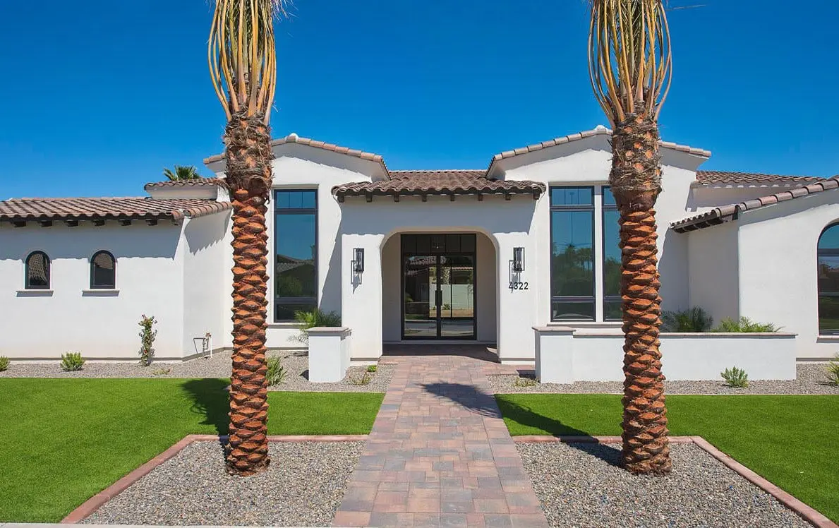 A modern, single-story white house with a tiled roof, large windows, and three palm trees in the front yard. A pathway made of interlocking bricks showcases expert paver installation leading to the front door. The outdoor space also features manicured lawns and gravel sections, fitting seamlessly into the charm of Surprise AZ.