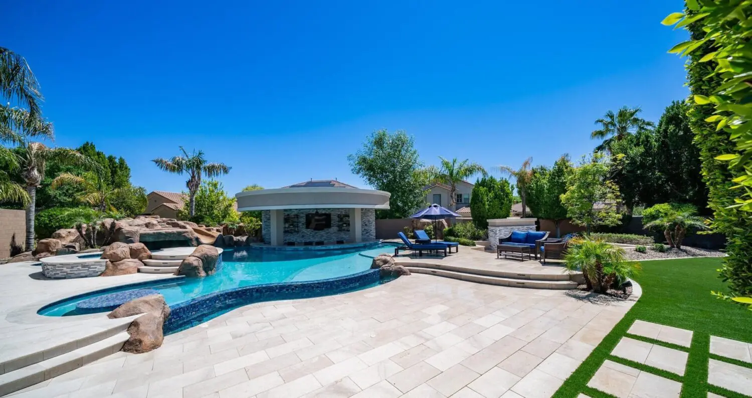 A luxurious backyard in Surprise, AZ features a pristine swimming pool with a rock waterfall, a hot tub, and a modern outdoor kitchen area. Lounge chairs and umbrellas are arranged on a spacious paver installation patio, surrounded by lush greenery and tall palm trees under a clear, blue sky.