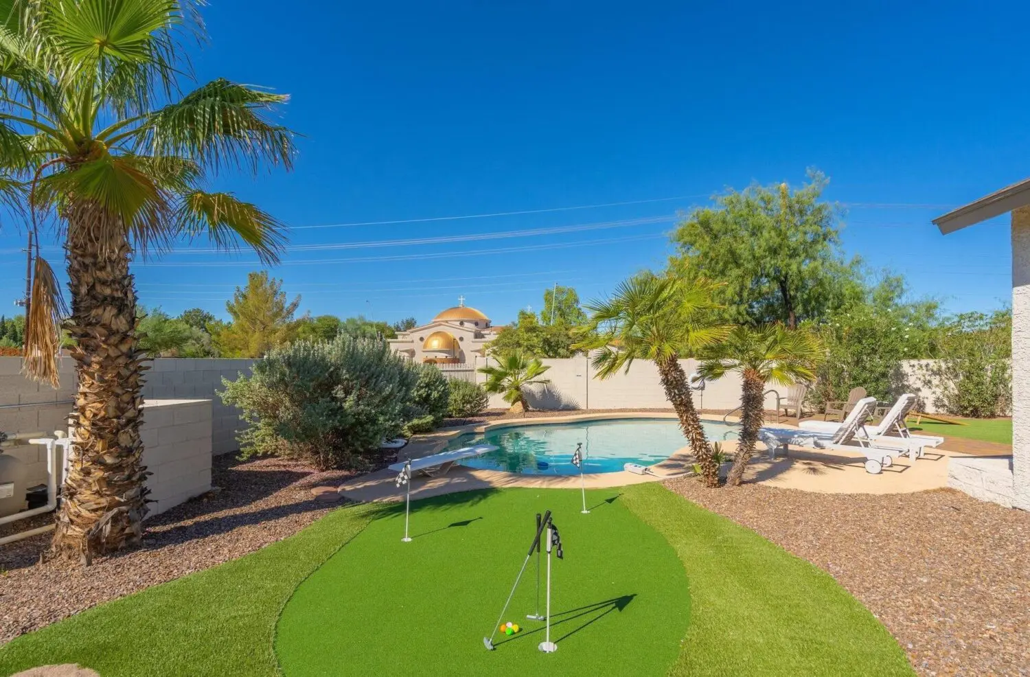 Backyard featuring a mini putting green with professional sports turf installation, surrounded by pebbled landscaping, various green plants, and palm trees. In the background, there's a kidney-shaped swimming pool with lounge chairs and a distant building with a dome against Arizona's bright, clear blue sky.