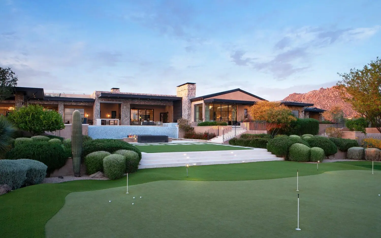 A spacious AZ backyard with a large artificial grass putting green. Multiple holes with flags and a chipping area on the fringe of the green. Steps leading up to a big house with artificial turf installed in front.