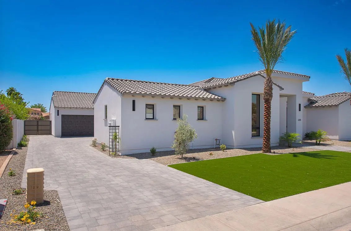 A modern white stucco house with a taupe tiled roof, located in the desert landscape of Surprise, AZ under clear blue skies. The home boasts a manicured front lawn, expert paver installation on the driveway leading to a garage, a few desert plants, and a tall palm tree enhancing the outdoor space.