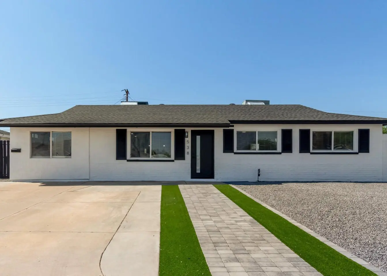 A single-story white house in Surprise, AZ, features a black front door and black window shutters. The front yard's outdoor space includes a concrete driveway on the left, a narrow strip of artificial grass leading up to the door, and a gravel area on the right. The sky is clear and blue.