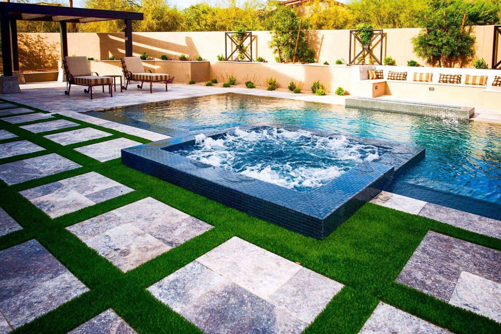 A modern backyard in Surprise, Arizona, features a tiled swimming pool and elevated hot tub, surrounded by neatly trimmed grass and stone pavers arranged in a geometric pattern. The area includes a shaded seating space with cushioned chairs and a beige privacy wall adorned with plants and residential synthetic turf.