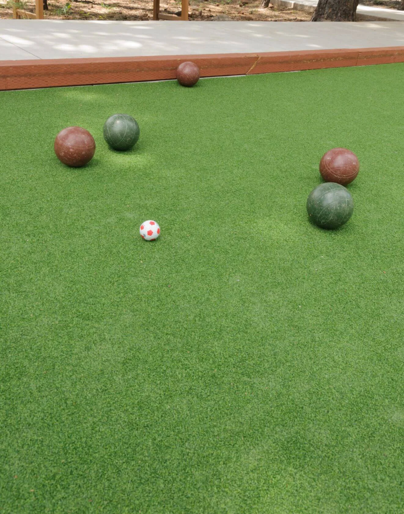 A bocce ball court with five bocce balls, including green and brown ones, positioned on professional sports turf installation. A tiny white ball with red dots, the pallino, is present in the middle of the synthetic turf-covered court. A wooden border frames part of the playing area in Arizona.