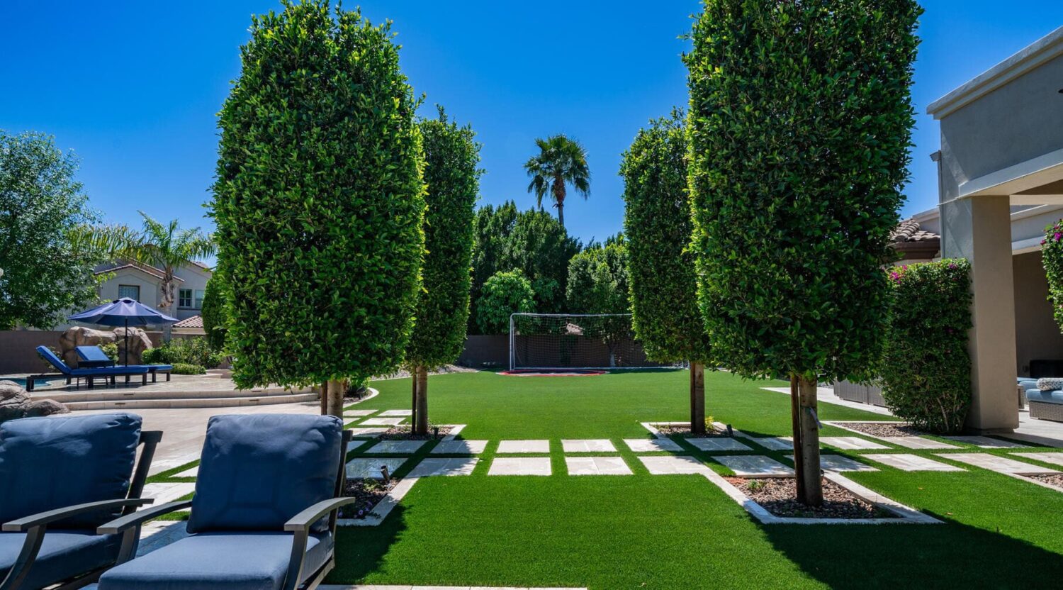 Synthetic turf in a yard with a paver path through the middle, and trees on either side. There is a soccer goal in the back for playing soccer on the sports turf section of the yard