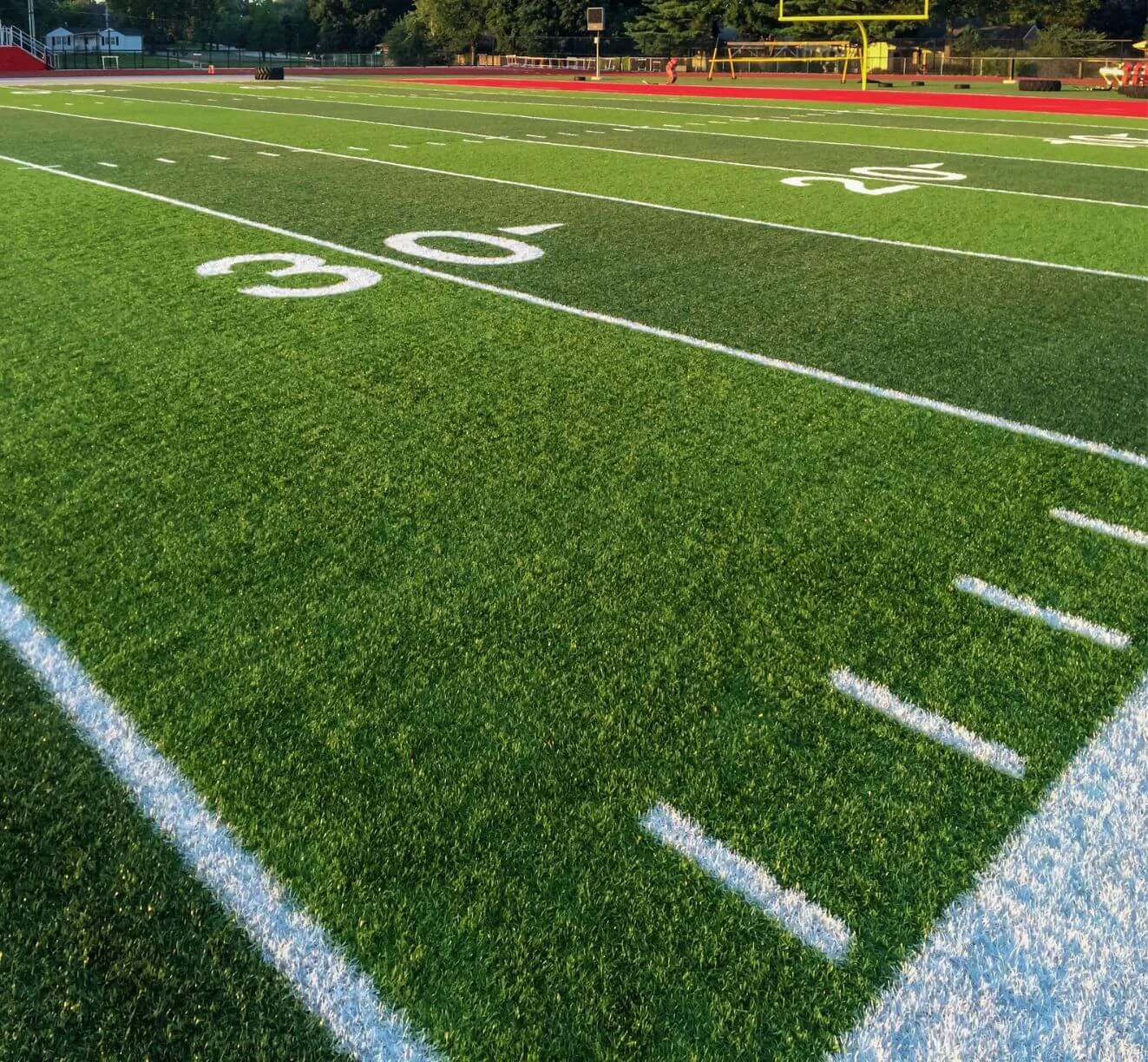 A green football field with white yard lines, focusing on the 30-yard line. The yellow goalposts are visible in the background against a setting sun and a backdrop of trees and a red athletic track, showcasing Arizona's top-tier professional sports turf installation.