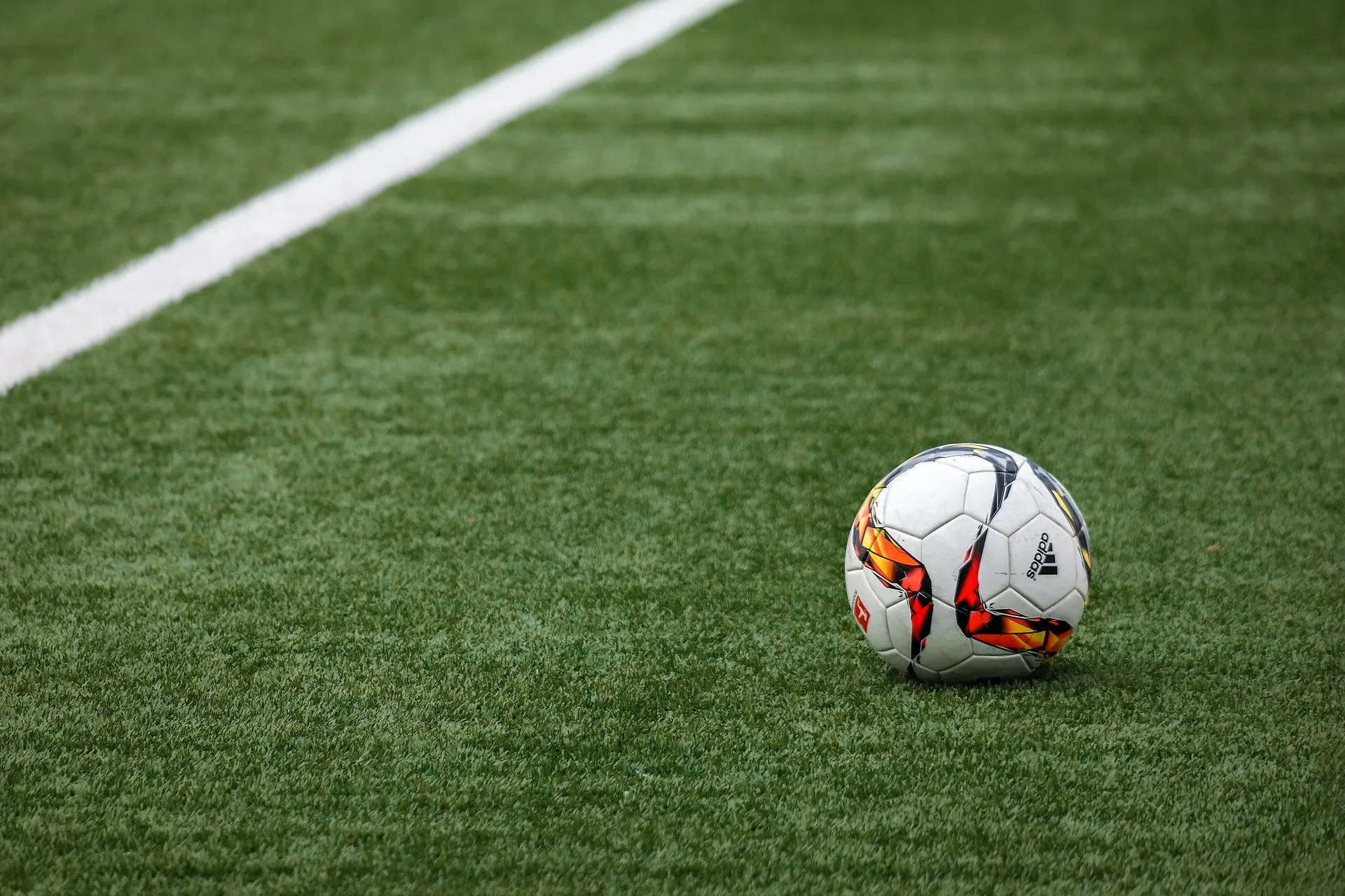 A white soccer ball with colorful design accents rests on a green synthetic turf field near a white boundary line. The vibrant texture of the grass contrasts with the detailed patterns on the ball.