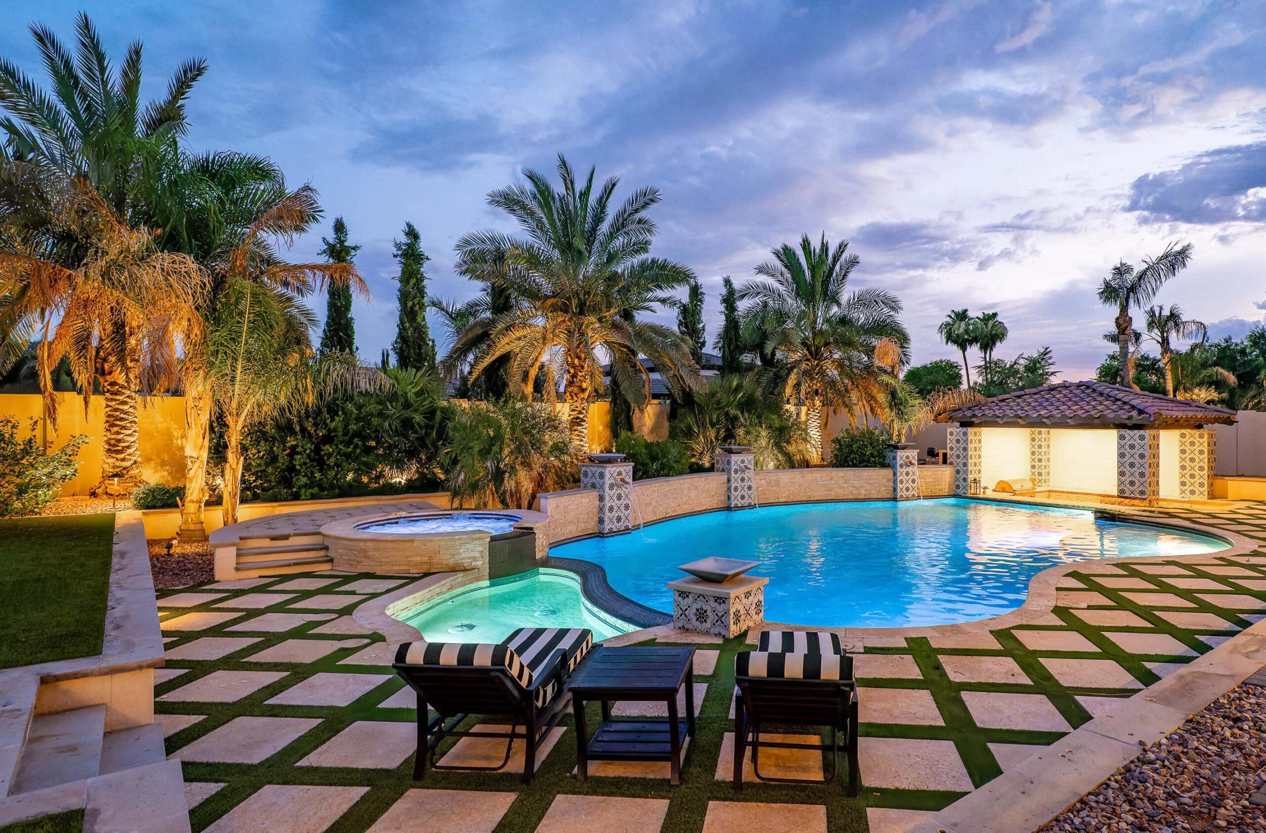 A luxurious backyard features a concrete pavers with artificial grass strips in between. A large, illuminated swimming pool with an attached hot tub, surrounded by palm trees. There are two lounge chairs with striped cushions and a small table on a patterned stone and artificial grass patio.